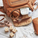 A Chloe - Caramel diaper bag with baby items and blocks on a wooden floor.