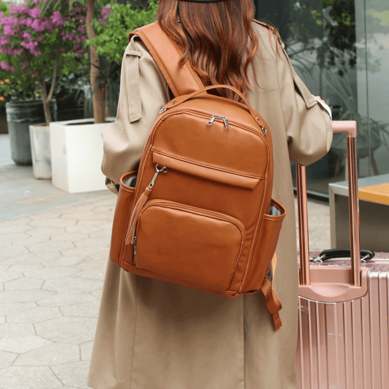 A woman in a trench coat is carrying the Olivia - Caramel backpack from Mother and Baby.
