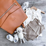A Scarlett - Caramel diaper bag with baby clothes and shoes on a wooden floor, by Mother and Baby.