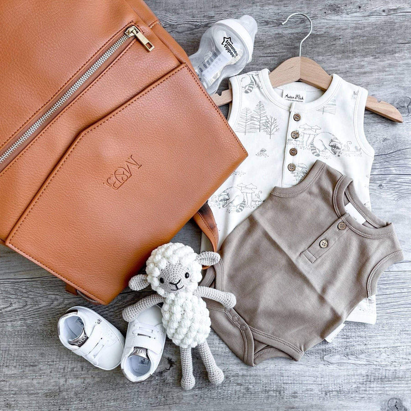 A Scarlett - Caramel diaper bag with baby clothes and shoes on a wooden floor, by Mother and Baby.